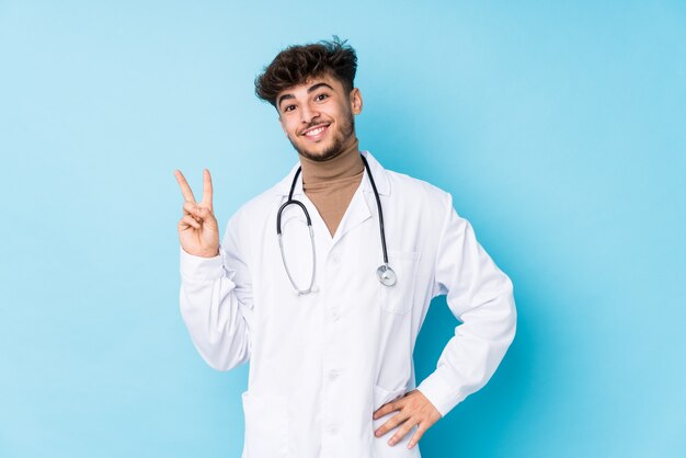 Young arab doctor man idolated joyful and carefree showing a peace symbol with fingers.