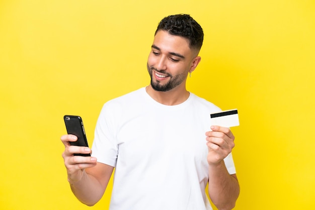 クレジット カードで携帯電話で購入する黄色の背景に分離された若いアラブのハンサムな男