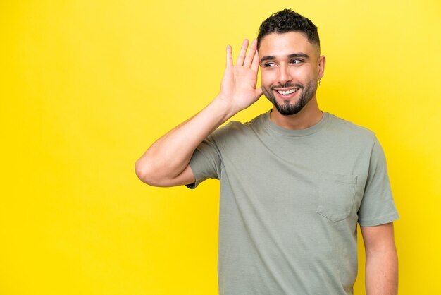 Young arab handsome man isolated on yellow background listening to something by putting hand on the ear