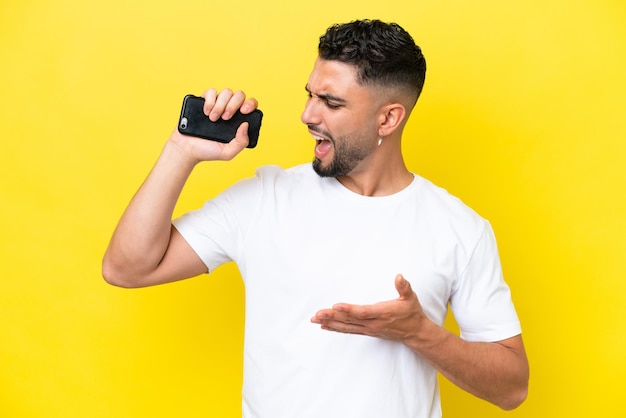 Young Arab handsome man isolated on yellow background using mobile phone and singing