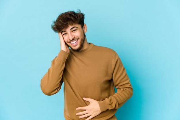 Young arab man on blue laughs happily and has fun keeping hands on stomach.