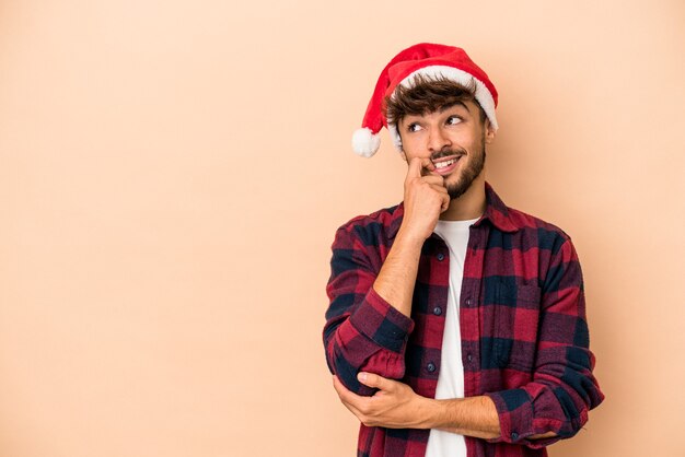 ベージュの背景に分離されたクリスマスを祝う若いアラブ人は、コピースペースを見ている何かについて考えてリラックスしました。