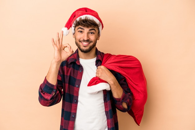 Giovane uomo arabo travestito da babbo natale isolato su sfondo beige allegro e fiducioso che mostra gesto ok.