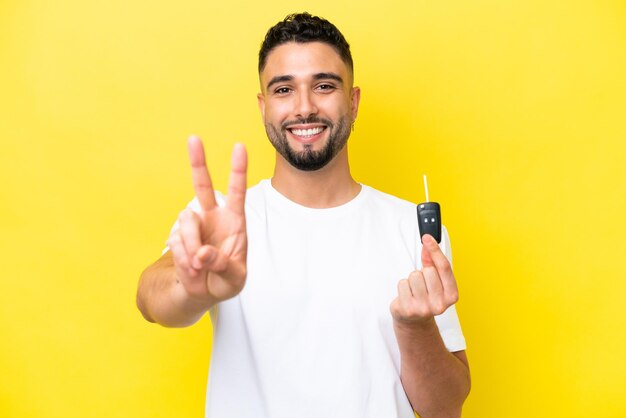 Young Arab man holding car keys isolated on yellow background smiling and showing victory sign