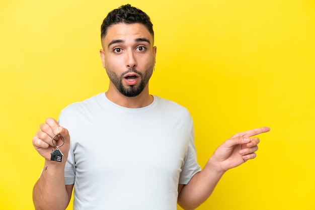 Young Arab man holding home keys isolated on yellow background surprised and pointing side