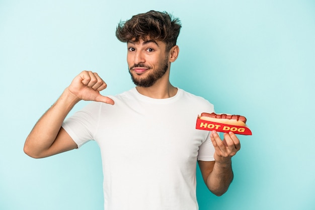 Young arab man holding a hotdog isolated on blue background feels proud and self confident, example to follow.