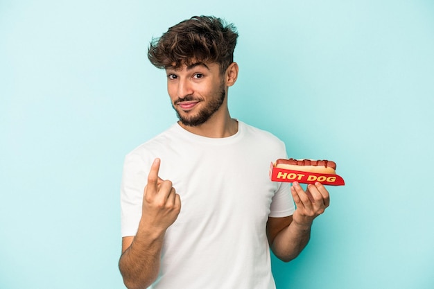 Young arab man holding a hotdog isolated on blue background pointing with finger at you as if inviting come closer.