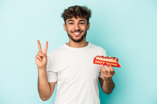 Giovane uomo arabo che tiene un hotdog isolato su sfondo blu che mostra il numero due con le dita.