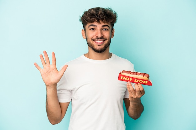 Young arab man holding a hotdog isolated on blue background smiling cheerful showing number five with fingers.