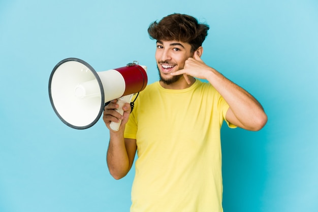 Giovane uomo arabo che tiene un megafono che mostra un gesto di chiamata di telefono cellulare con le dita.