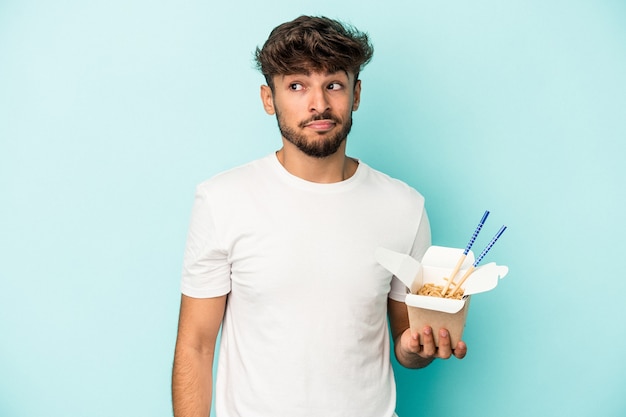 Young arab man holding a take away noodles isolated on blue background confused, feels doubtful and unsure.