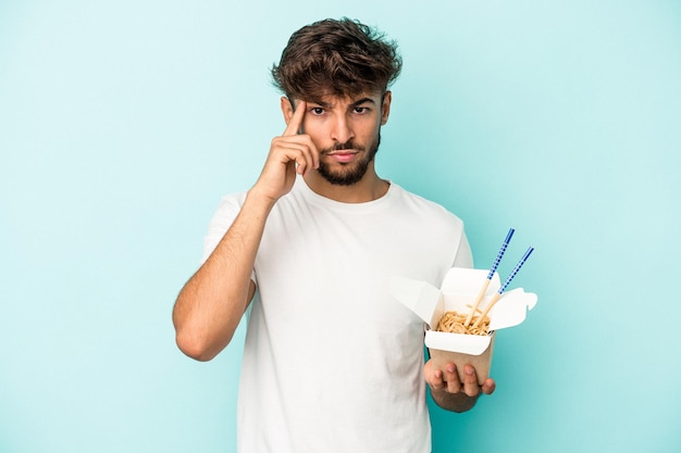 Young arab man holding a take away noodles isolated on blue background pointing temple with finger, thinking, focused on a task.