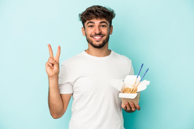 Young arab man holding a take away noodles isolated on blue background showing number two with fingers.