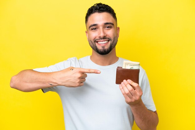 Young Arab man holding a wallet isolated on yellow background and pointing it