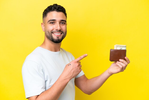 Young Arab man holding a wallet isolated on yellow background and pointing it