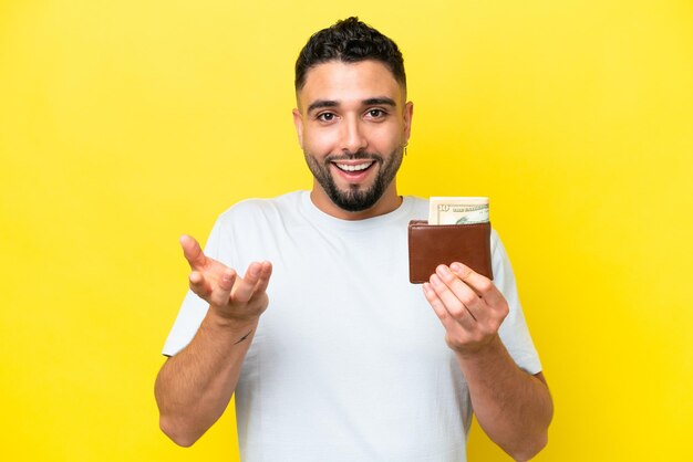 Young Arab man holding a wallet isolated on yellow background with shocked facial expression