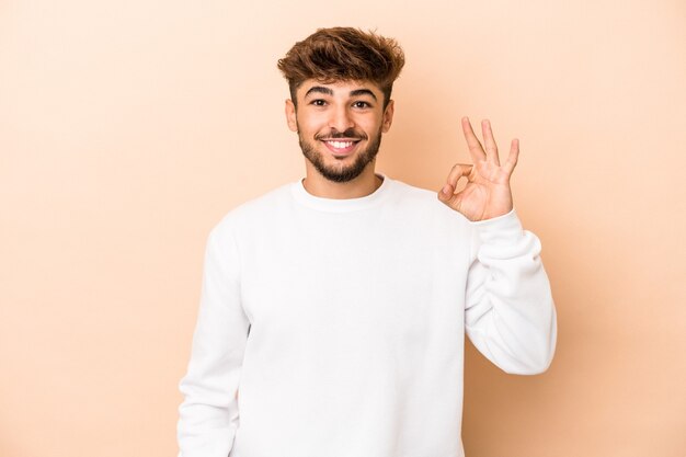 Young arab man isolated on beige background cheerful and confident showing ok gesture.