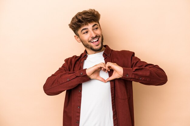 Young arab man isolated on beige background smiling and showing a heart shape with hands.