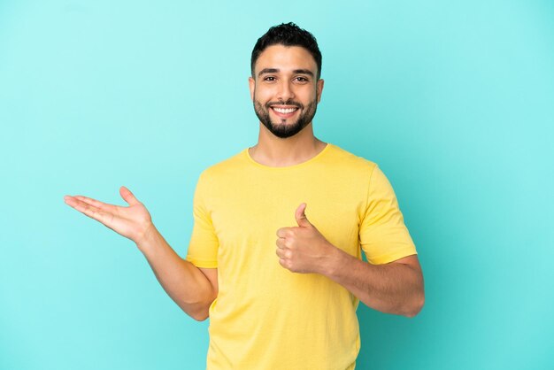 Young arab man isolated on blue background holding copyspace imaginary on the palm to insert an ad and with thumbs up