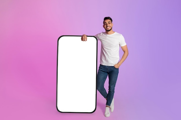 Young arab man leaning on big cellphone with empty screen offering space for your ad mobile app or