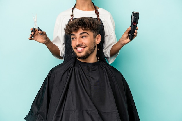 Young arab man ready to get a haircut isolated on blue background looks aside smiling, cheerful and pleasant.