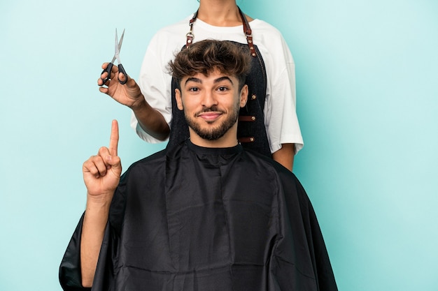 Giovane uomo arabo pronto a ottenere un taglio di capelli isolato su sfondo blu che mostra il numero uno con il dito.