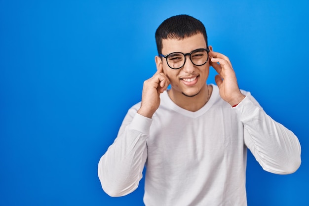 Young arab man wearing casual white shirt and glasses covering ears with fingers with annoyed expression for the noise of loud music deaf concept