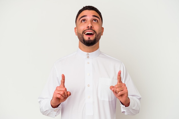 Young arab man wearing typical arab clothes isolated on white wall pointing upside with opened mouth.