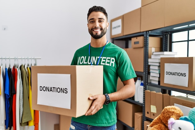 Young arab man wearing volunteer uniform holding donations box at charity center