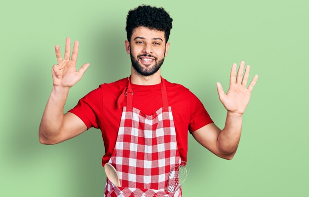 調理用エプロンを着たひげを生やした若いアラブ人男性が、自信と幸せな笑みを浮かべながら、指番号9を示して上を向いている