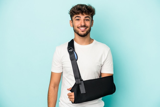 Young arab man with broken hand isolated on blue background happy, smiling and cheerful.