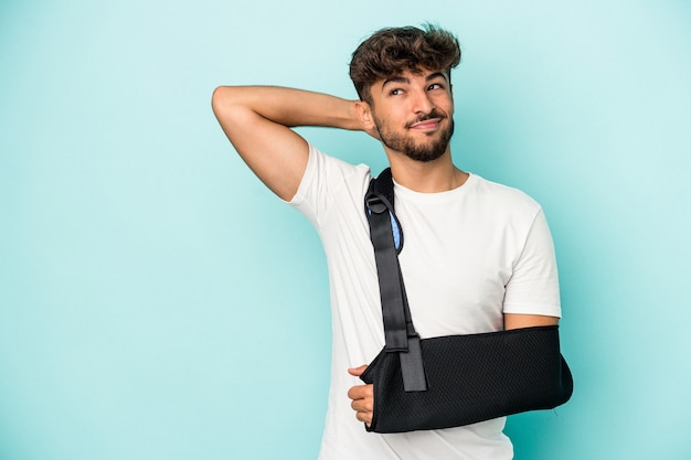 Young arab man with broken hand isolated on blue background touching back of head, thinking and making a choice.