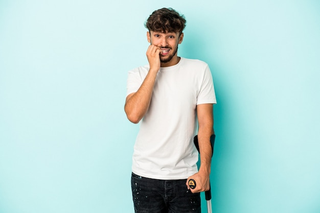 Young arab man with crutches isolated on blue background biting fingernails, nervous and very anxious.