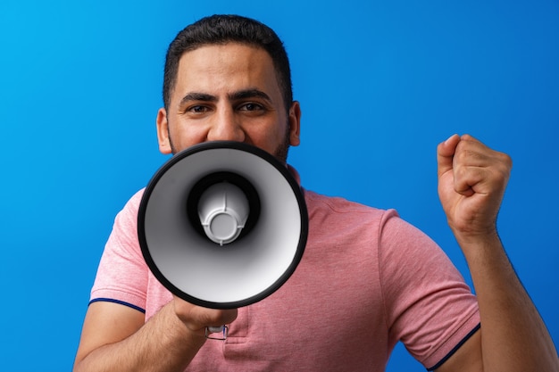 Young arab man with megaphone proclaiming something isolated on blue background