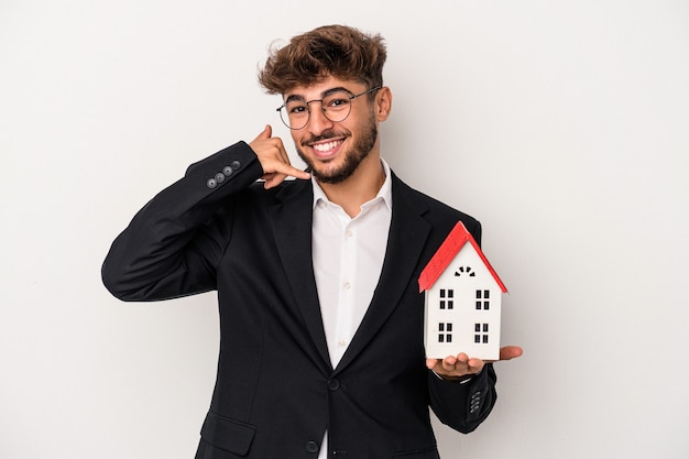 Young arab real estate man holding a model house isolated on isolated background showing a mobile phone call gesture with fingers.