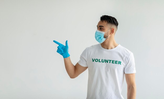 Young arab volunteer in medical mask and gloves pointing and looking aside at empty space over light wall panorama