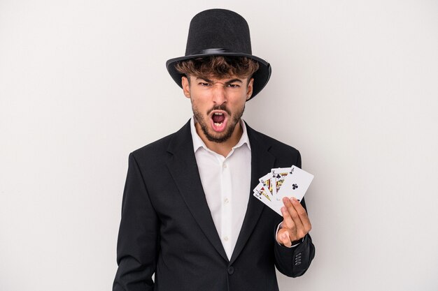 Young arab wizard man holding a magic cards isolated on white background screaming very angry and aggressive.