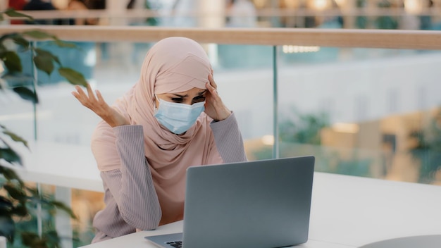 Young arab woman in hijab wearing protective mask reading email on laptop receiving bad news girl