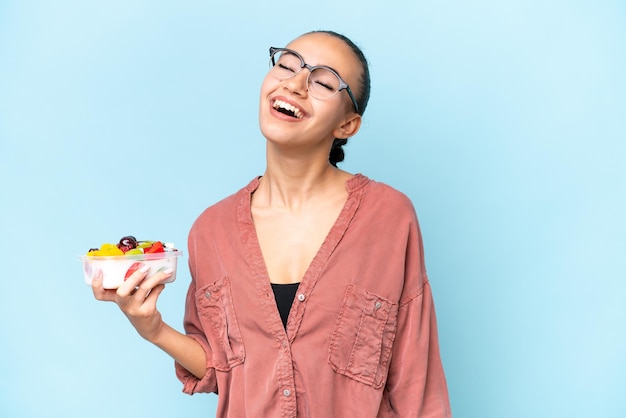 Young Arab woman holding a bowl of fruit isolated on blue background laughing