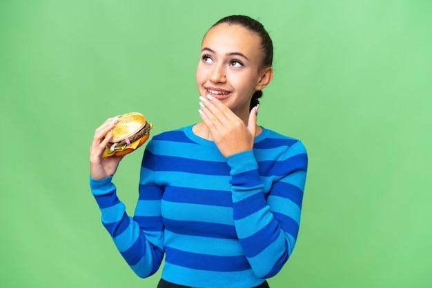 Young Arab woman holding a burger over isolated background looking up while smiling