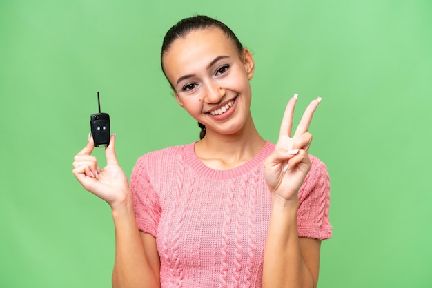 Young Arab woman holding car keys over isolated background smiling and showing victory sign