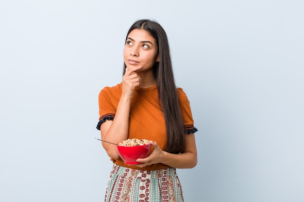 Young arab woman holding a cereal bowl looking sideways with doubtful and skeptical expression.