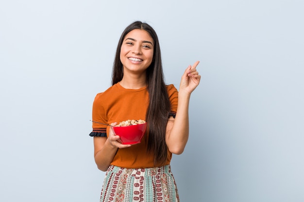 Young arab woman holding a cereal bowl smiling cheerfully pointing with forefinger away.