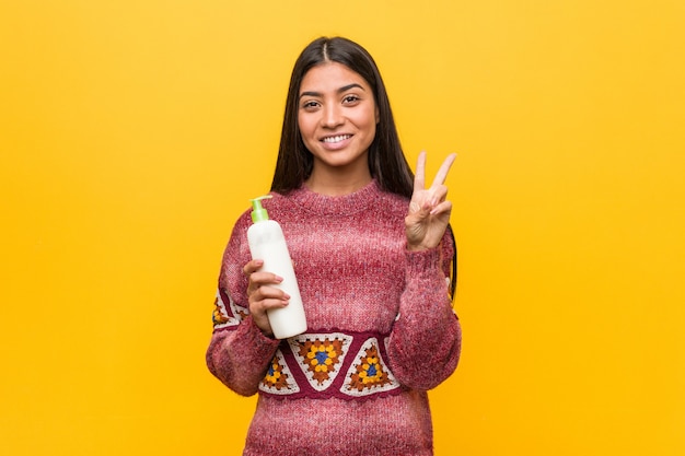 Young arab woman holding a cream bottle showing number two with fingers.