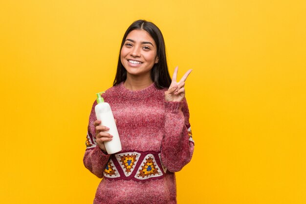 Young arab woman holding a cream bottle showing victory sign and smiling broadly.