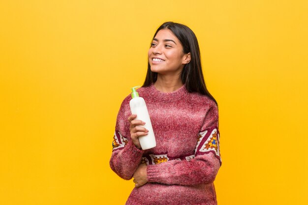 Giovane donna araba che tiene una bottiglia di crema sorridente fiducioso con le braccia incrociate.