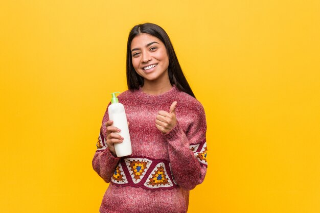 Young arab woman holding a cream bottle smiling and raising thumb up