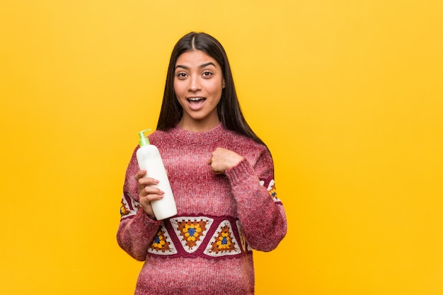 Young arab woman holding a cream bottle surprised pointing at himself, smiling broadly.