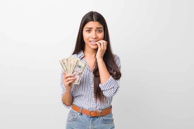 Young arab woman holding dollars biting fingernails, nervous and very anxious.