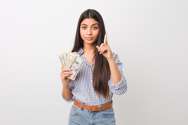 Young arab woman holding dollars showing number one with finger.